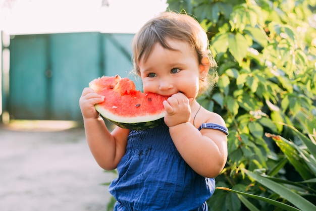 Encantadora niña comiendo sandía en el patio en el fondo de las plantas, muy lindo