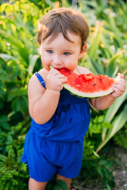 Encantadora niña comiendo sandía en el patio en el fondo de las plantas, muy lindo