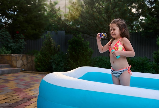 Encantadora niña pequeña en traje de baño se divierte en la piscina inflable sopla burbujas de jabón al atardecer