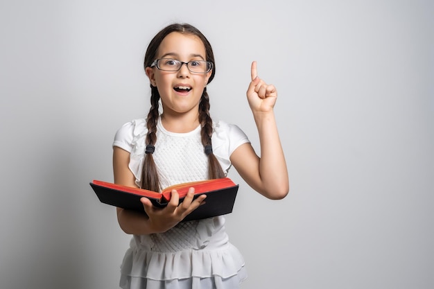 Encantadora niña de pie y sosteniendo un libro sobre fondo blanco.