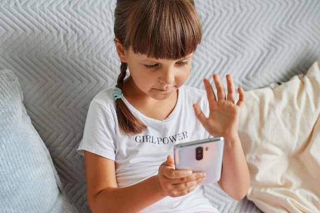 Encantadora niña sentada en el sofá con teléfono móvil y tener videollamadas con amigos, mirando la pantalla del dispositivo y agitando la mano a la cámara del dispositivo, niño con ropa informal.