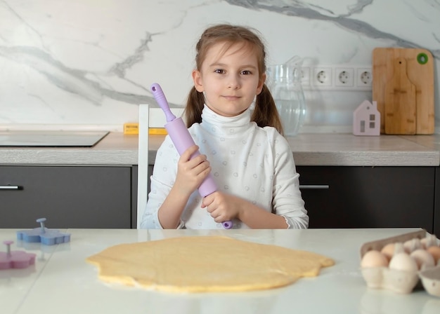 Foto una encantadora niña de siete años está aprendiendo a cocinar en una cocina blanca. un niño feliz cocina, hace masa, hornea galletas. un pequeño ayudante ayuda en la cocina.