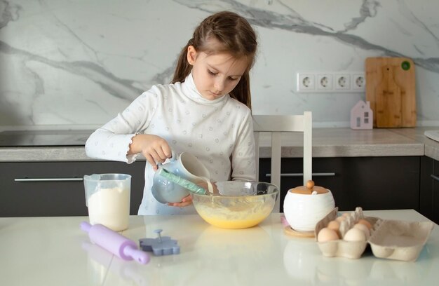 Una encantadora niña de siete años está aprendiendo a cocinar en una cocina blanca. Un niño feliz cocina, hace masa, hornea galletas. Un pequeño ayudante ayuda en la cocina.