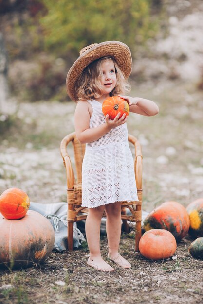 encantadora niña en un sombrero de paja, silla de mimbre, calabazas, otoño