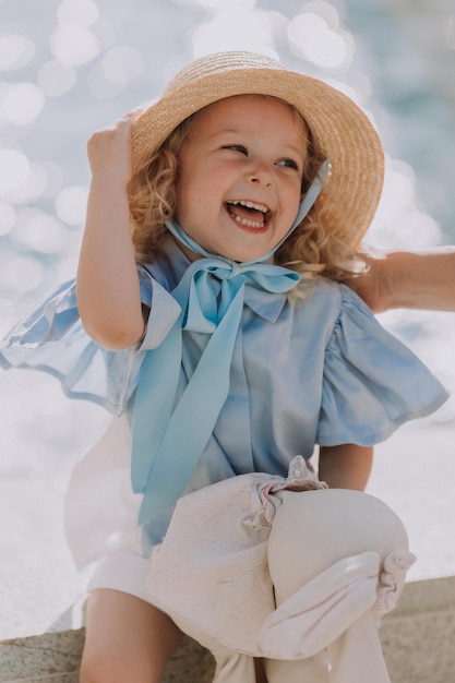 encantadora niñita rubia con sombrero de paja y vestido azul juega con un conejo de peluche al aire libre