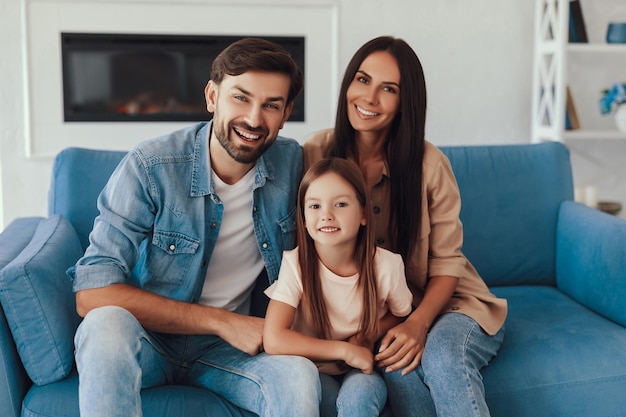 Encantadora niñita sonriendo mientras se sienta entre su joven madre y su padre en un acogedor sofá azul