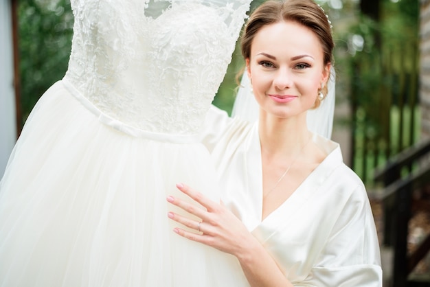 Foto encantadora novia con cabello rubio oscuro sostiene su vestido delicadamente de pie afuera