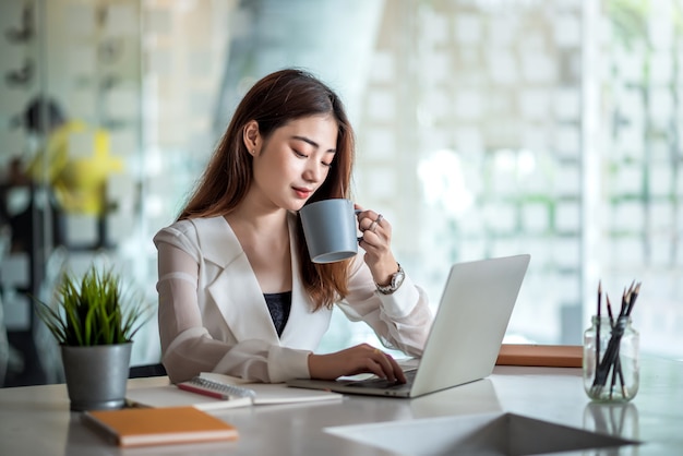 Encantadora oficinista asiática trabajando en una computadora portátil y disfrutando de beber café en una oficina moderna