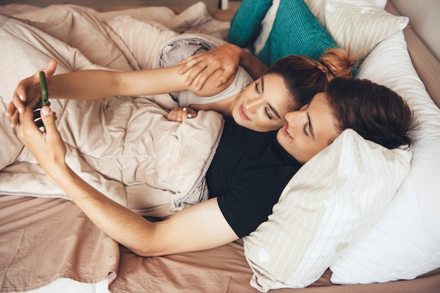 Encantadora pareja acostada en la cama y sonríe mientras hace un selfie con un teléfono móvil