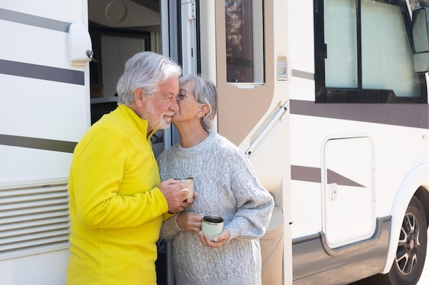 Encantadora pareja de ancianos besándose en la puerta de su autocaravana mientras beben un café Sonrientes ancianos atractivos disfrutando de la jubilación en libertad viajes de vacaciones en el bosque