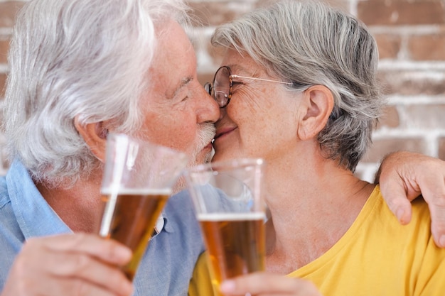 Encantadora pareja de ancianos brindando con un vaso de cerveza rubia mientras se besan
