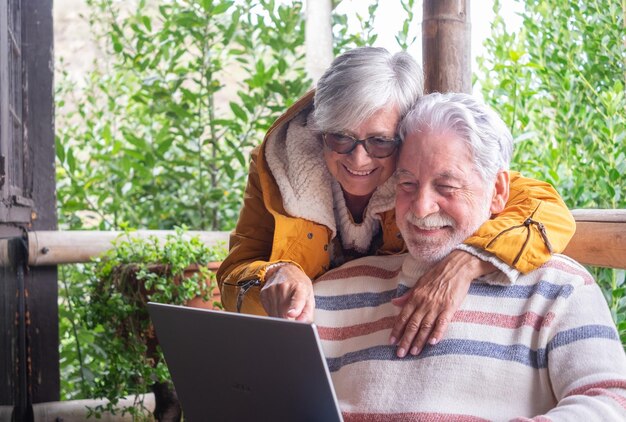 Una encantadora pareja de ancianos de cabello blanco se abrazan en el balcón de un chalet de montaña mientras el hombre usa una computadora portátil