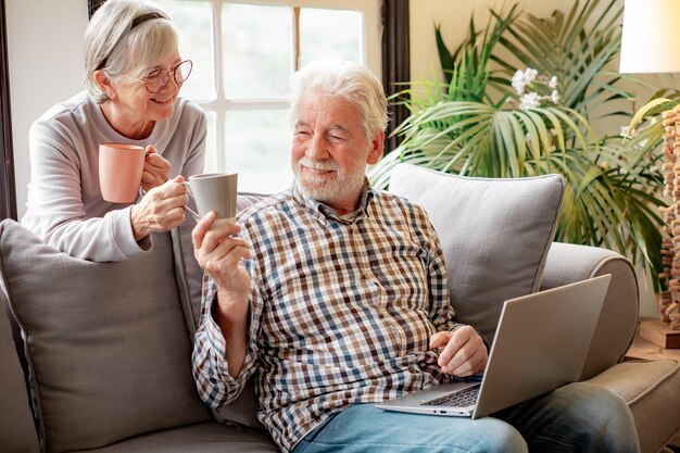 Encantadora pareja de ancianos relajándose en el sofá en la sala de estar bebiendo una taza de té disfrutando de la jubilación