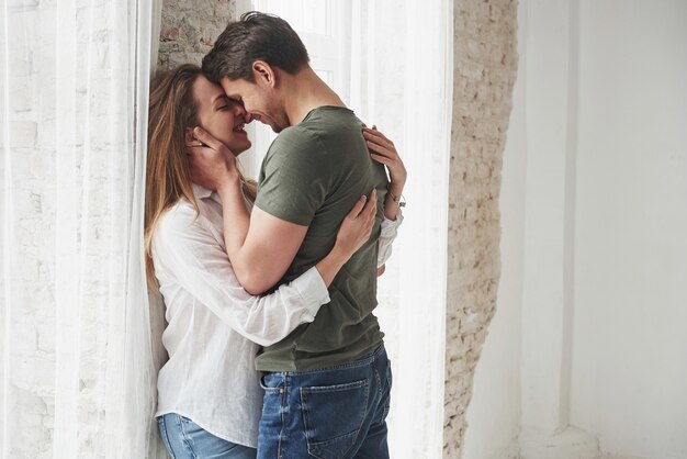 Encantadora pareja besándose y disfrutando cerca de la ventana de su nuevo hogar.