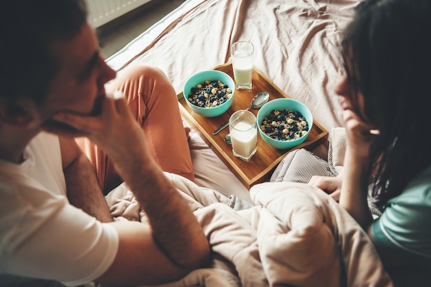 Encantadora pareja en la cama mirándose antes de comer cereales con leche por la mañana