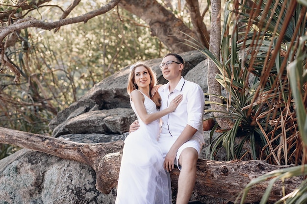 Encantadora pareja casada feliz, santificada por el sol, sentada en un tronco de árbol en la jungla y mirando a lo lejos, sonriendo alegremente. concepto de amor