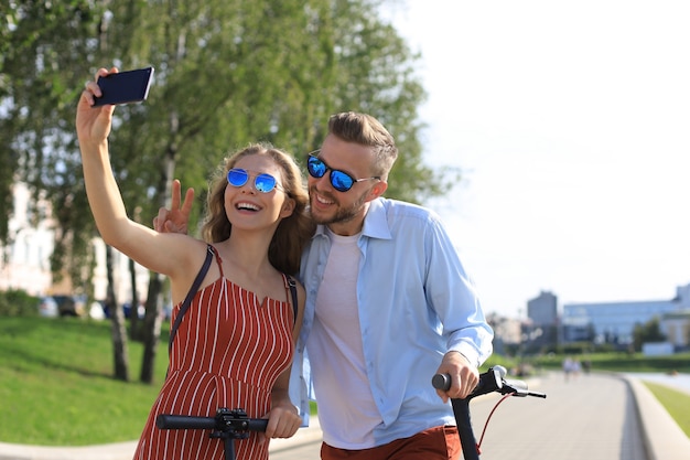 Encantadora pareja divirtiéndose conduciendo scooter eléctrico por la ciudad, tomando un descanso, tomando selfie.
