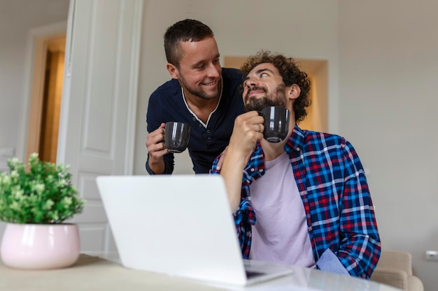 Encantadora pareja gay riendo juntos mientras están sentados en su sala de estar en casa Dos románticos jóvenes amantes divirtiéndose navegando por internet en el interior Joven pareja gay viviendo juntos