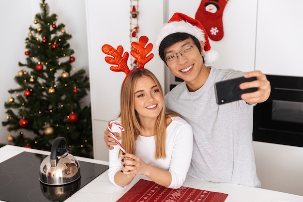 Encantadora pareja, hombre y mujer con traje de Navidad, de pie en la cocina luminosa y tomando una foto selfie en el teléfono móvil