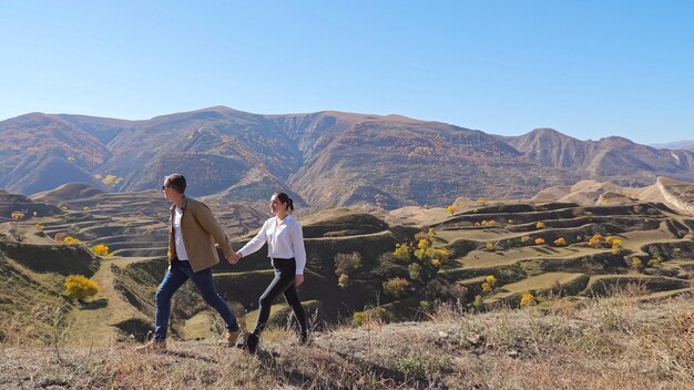 Encantadora pareja joven corre a lo largo de la cresta de la colina con pasto seco contra la pendiente de la cascada con raros árboles amarillos en Highland en un bonito día soleado