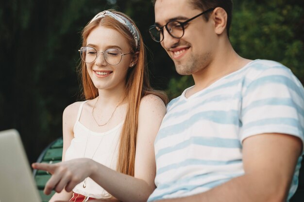 Encantadora pareja joven mirando su portátil sonriendo mientras trabaja independientemente al aire libre en el parque en una playa. Pareja joven en programa que trabaja afuera.