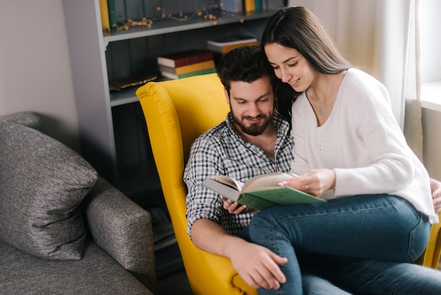 Encantadora pareja joven positiva leyendo un libro juntos sentados en una silla amarilla