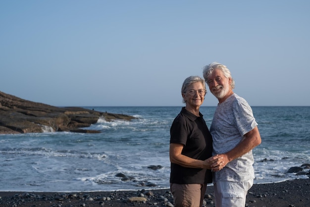 Encantadora pareja mayor caucásica de pie en la playa al atardecer mirando a la cámara jubilados sonrientes disfrutando del tiempo libre vacaciones en el mar o la jubilación