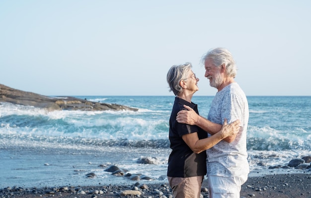 Encantadora pareja mayor caucásica de pie en la playa al atardecer mirándose a los ojos jubilados sonrientes disfrutando del tiempo libre vacaciones en el mar o jubilación