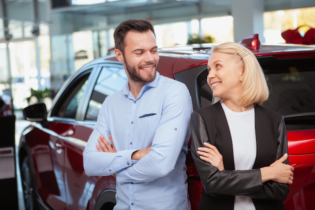 Encantadora pareja sonriendo alegremente el uno al otro, de pie cerca de su auto nuevo en el concesionario