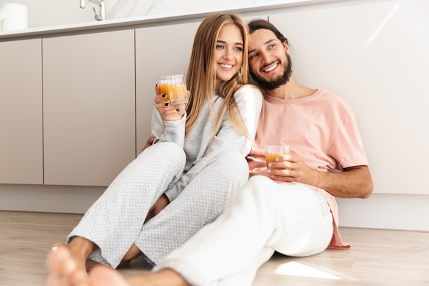 Encantadora pareja sonriente abrazándose mientras está sentado en el piso de la cocina, bebiendo jugo de naranja