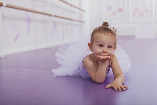 Encantadora pequeña bailarina soplando besos a la cámara, tirada en el suelo en la clase de ballet