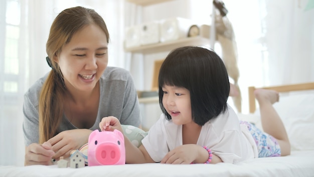 Encantadora pequeña niña asiática y madre poniendo dinero en la hucha para el ahorro