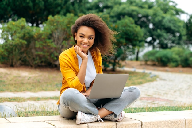 encantadora positiva inteligente afroamericana de cabello rizado chica del milenio estudiante independiente con ropa casual elegante sentada con una computadora portátil al aire libre mirando a la cámara sonriendo felizmente