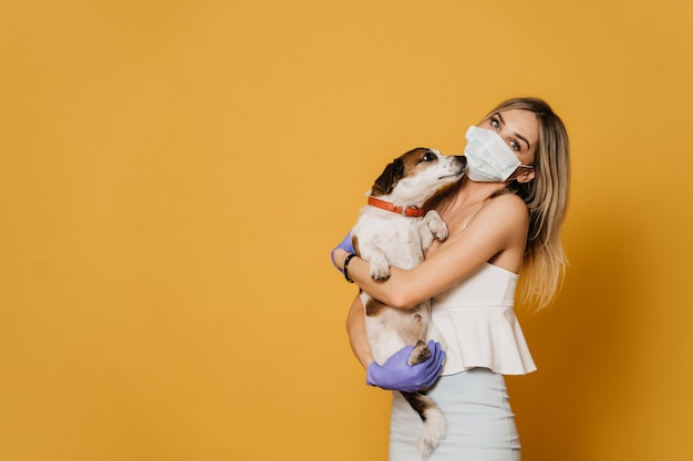 Encantadora rubia con máscara quirúrgica y guantes protectores, vestida con una blusa ligera y una falda abraza a su perro, cuida a tus amigos durante la cuarentena debido a la pandemia de coronavirus. Concepto Covid-19.