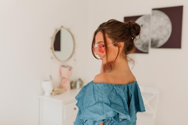 Encantadora senhora elegante com cabelos escuros usando óculos redondos e camiseta azul posando de trás enquanto olha para baixo sobre fundo claro