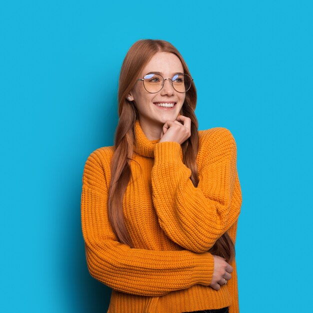 Encantadora senhora ruiva com sardas e óculos, um suéter de malha amarelo posando em uma parede azul