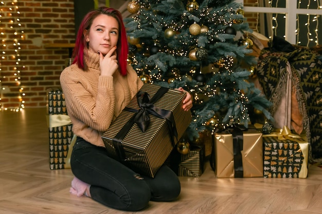 Encantadora señorita pensativa en un suéter con un regalo en sus manos en el suelo con el telón de fondo de un árbol de Navidad con regalos