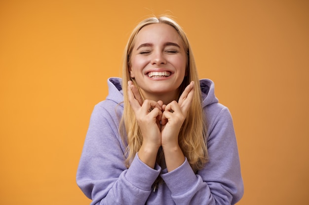 Encantadora sorte fofa jovem feliz esperançosa loira fecha os olhos sorrindo dedos cruzados positivos boa sorte quer ganhar fielmente orando fazer desejo desejo sonho tornado realidade, fundo laranja de pé.