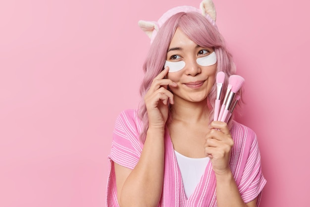 Encantadora y tierna mujer joven con cabello teñido de rosa aplica parches de belleza debajo de los ojos sostiene cepillos cosméticos que van a maquillarse tiene una expresión pensativa aislada sobre el área de espacio de copia de fondo rosado