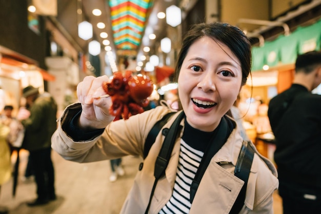 Encantadora turista parada en medio del mercado, frente a la cámara y mostrando el pequeño pulpo japonés. comida callejera tradicional en Japón. Vacaciones de viaje de Asia en Kioto.