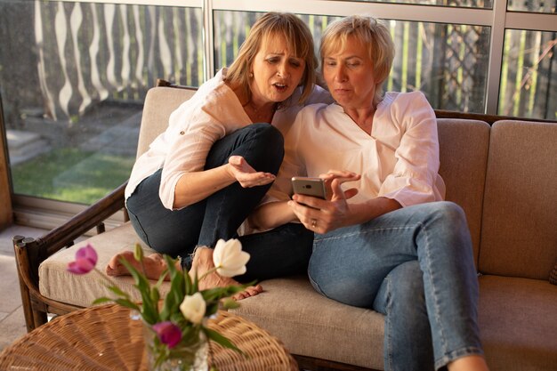 Encantadoras damas de 55 años usan teléfonos inteligentes para discutir la moda femenina, sentadas en el sofá a la mesa con flores en el patio de una casa de madera