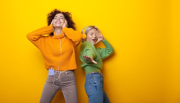 Encantadoras dos mujeres bailando en una pared amarilla con espacio libre escuchando música a través de auriculares