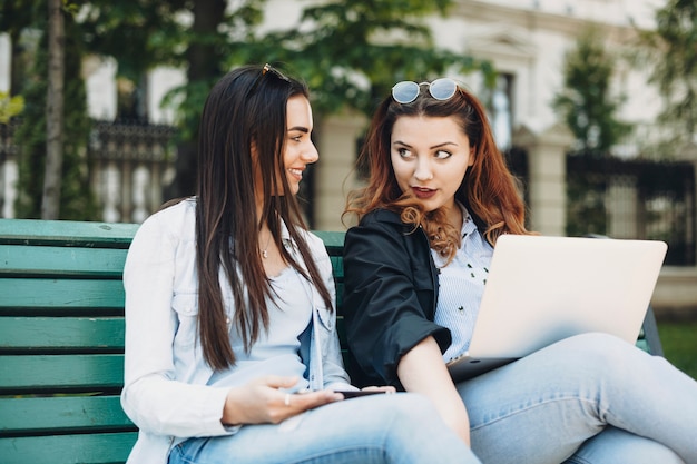 Encantadoras mujeres de talla grande hablando de algo interesante con su novia mientras están sentadas en un banco afuera con una computadora portátil en las piernas.