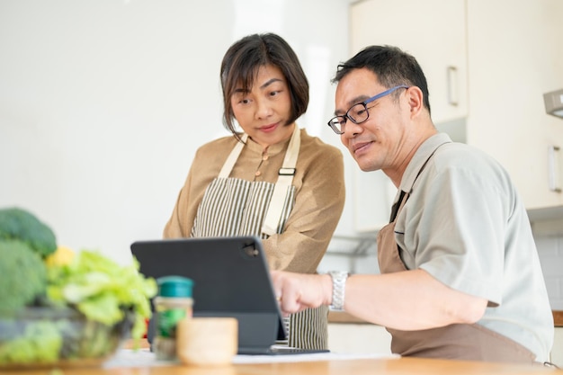 Unas encantadoras parejas asiáticas están mirando una receta en línea en una tableta y cocinando en la cocina juntos.