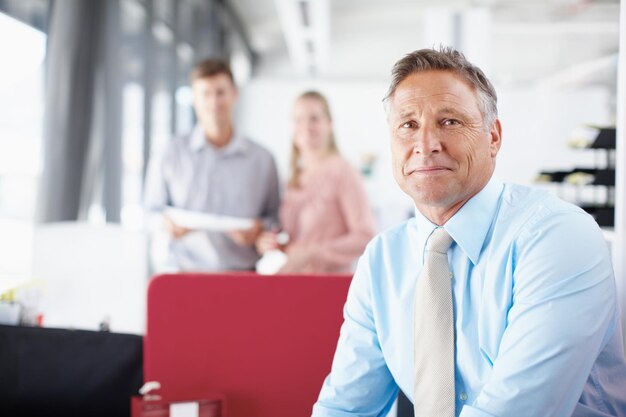 Le encantan los desafíos que le plantean los negocios Retrato de un gerente de negocios sénior sonriendo mientras sus colegas trabajan en segundo plano