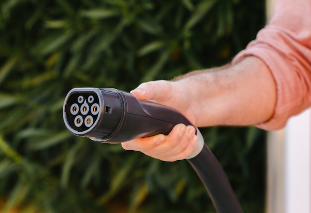 Enchufe de coche eléctrico sostenido por la mano de un hombre con fondo verde natural. Concepto de movilidad ecológica