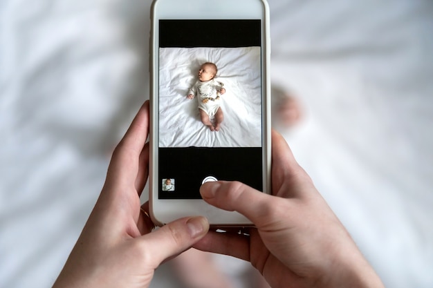 Por encima de un alto ángulo de disparo de la feliz mamá tomando fotos a su bebé o niña en la cama con un teléfono inteligente en casa