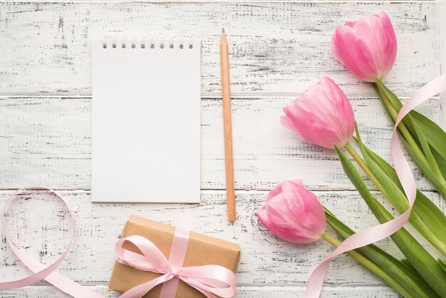 Encima de la foto plana superior de tulipanes de color pastel sobre tallos verdes envueltos en una caja de regalo con cinta y cuaderno con una página en blanco de papel para maquetas y lápiz sobre una mesa de madera