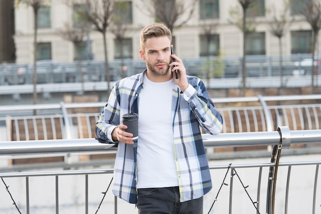 Encontrémonos en unos minutos. Comunicación de la vida moderna. Chico moderno con antecedentes urbanos de teléfonos inteligentes. Guapo teléfono móvil y taza de café. Tecnología moderna. Conversación con un amigo. Llámame luego.