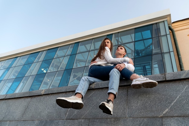 Encontro de um jovem casal. A menina está sentada no colo do cara. Edifício moderno da cidade ao fundo. Vista de baixo.